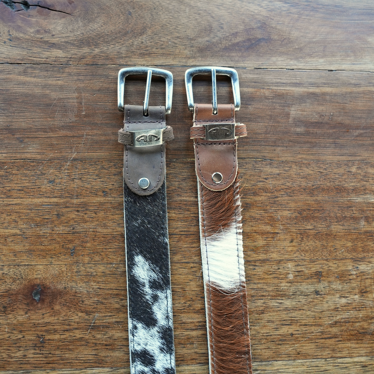 Two leather belts with buckles laid flat on a wooden surface. One belt has a black-and-white cowhide pattern, and the other has a brown-and-white cowhide pattern.