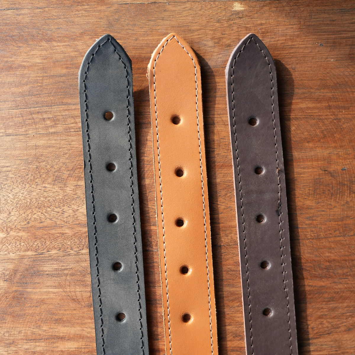 Three leather belts in black, tan, and dark brown, laid flat side-by-side on a wooden surface showing the belt ends.