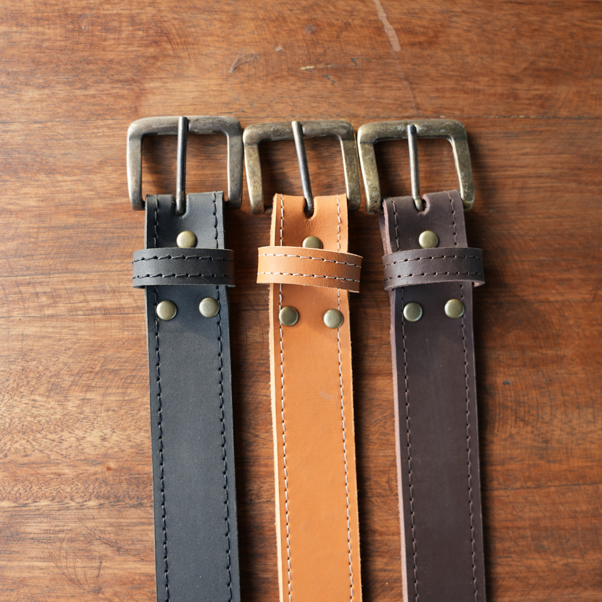 Three leather belts in black, tan, and dark brown, laid flat on a rustic wooden surface. Each belt features a brass buckle, a matching keeper loop, and brass rivets.