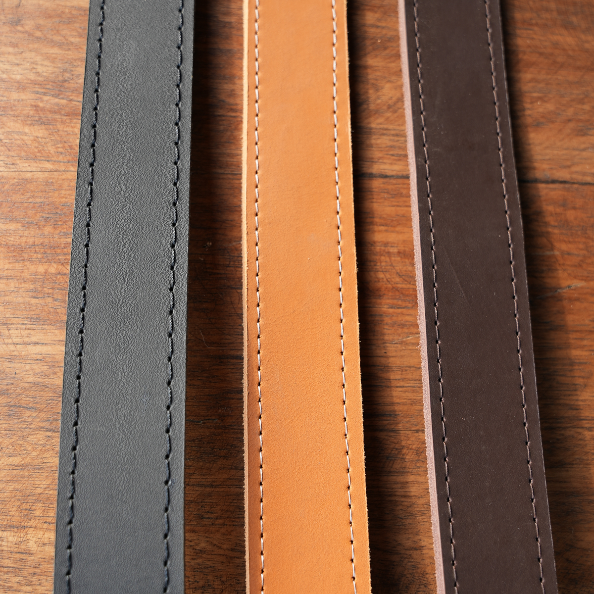 Three leather belts in black, tan, and brown, displayed vertically side-by-side on a wooden surface.