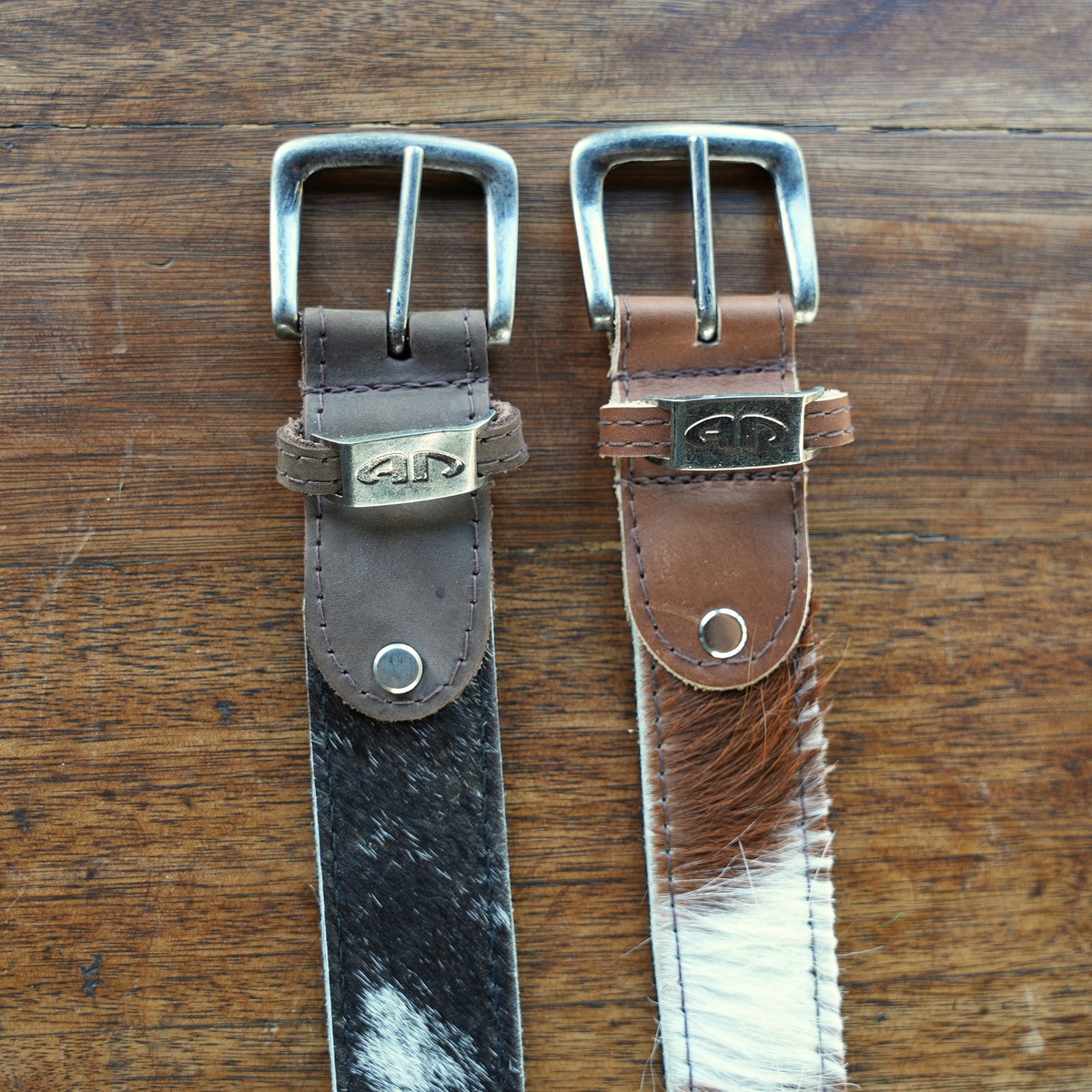 Two hide and leather belts displayed side by side on a wooden surface. The left belt is dark brown with a black-and-white cowhide pattern, and the right belt is medium brown with a white-and-brown cowhide pattern.