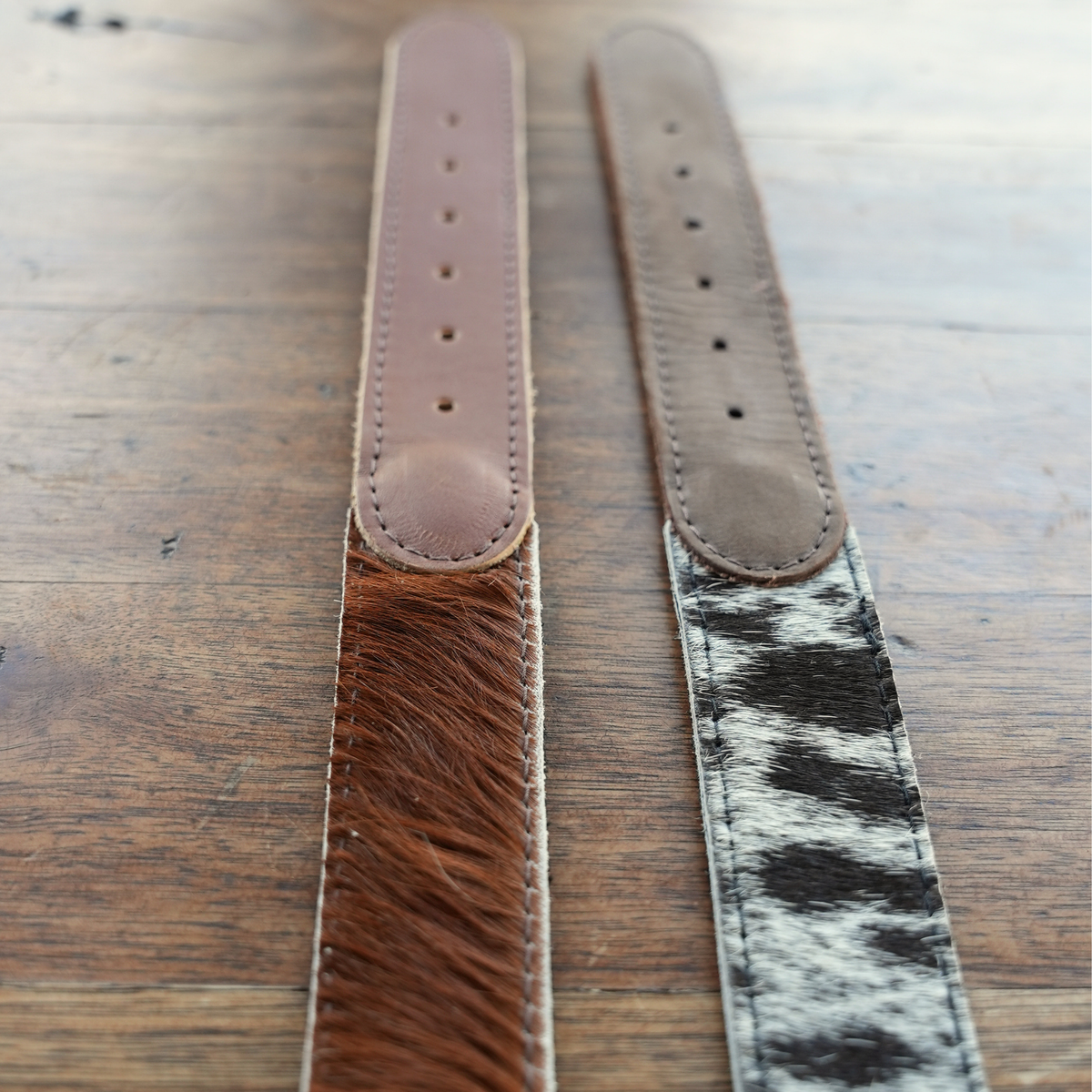 Two belt ends laid flat on a wooden surface, one with brown hair-on-hide and the other with black-and-white cowhide, both with brown leather end sections.