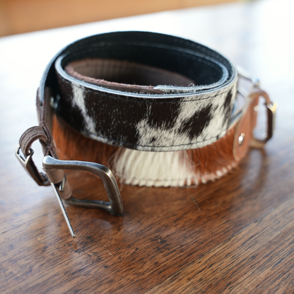 Two western-style belts with black, white and brown cowhide resting on a wooden surface.