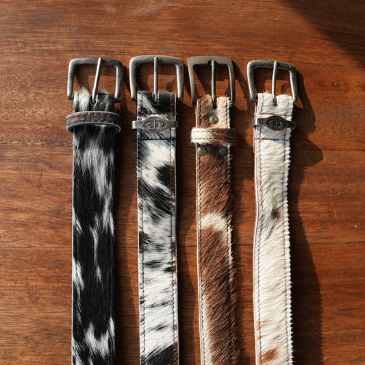 Four nguni cowhide belts with different patterns and metal buckles, displayed vertically on a wooden surface.