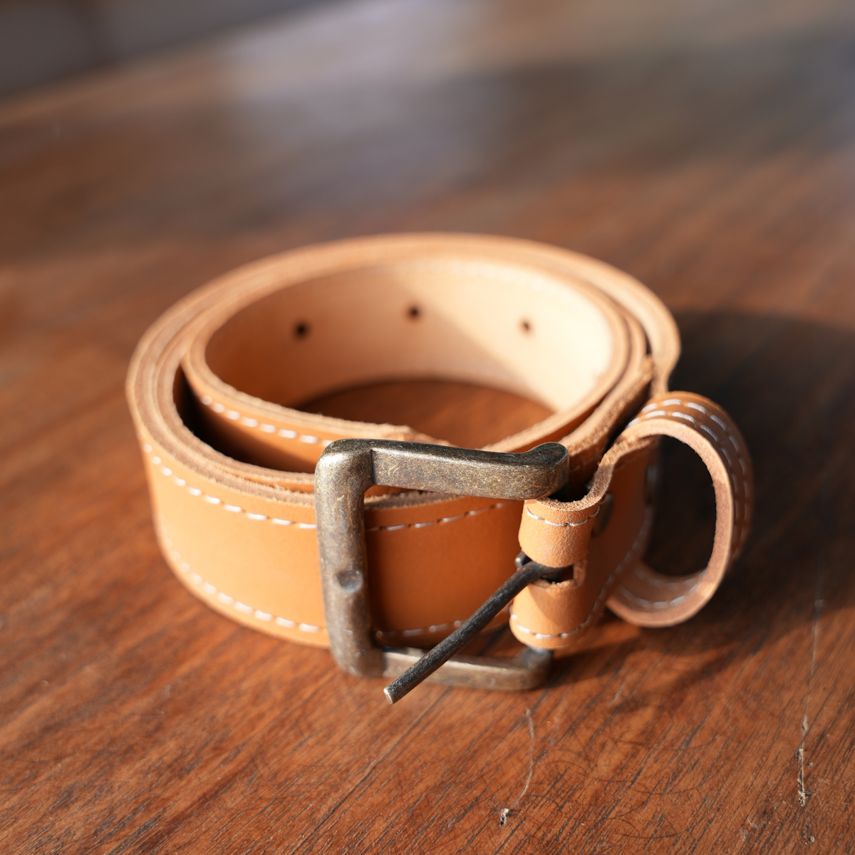 A light tan leather belt with a metal buckle, coiled on a wooden surface.